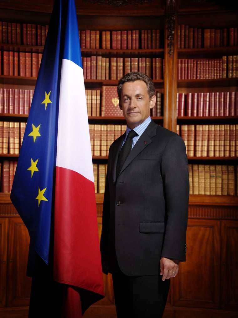 Vue officielle du Président de la République Nicolas Sarkozy posant aux côtés des drapeaux européen et français, devant une bibliothèque pleine de livres anciens.