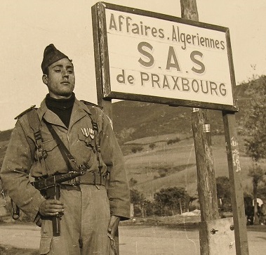 Un harki en tenue militaire avec son pistolet mitrailleur (PM MAT 49) posant à côté de la pancarte annonçant "Affaires Algériennes, section administrative spécialisée (SAS) de Praxbourg" [1958-1959]